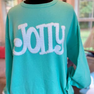 Puede incluir: Una sudadera turquesa con la palabra "JOLLY" en letras grandes, blancas y difusas en la parte delantera. La sudadera se muestra en un maniquí negro y tiene mangas largas.
