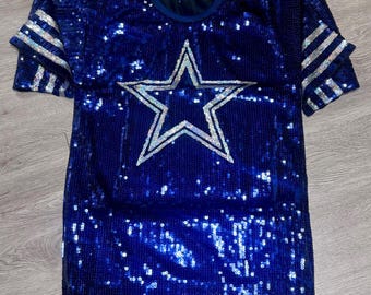 May include: A royal blue sequined shirt with a large silver star in the center. The sleeves have silver stripes. The shirt has a round neckline and is made of shiny material.
