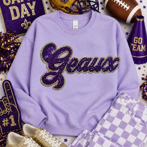 May include: A light purple crewneck sweatshirt featuring the word Geaux in purple sequins with a gold sequin border. The garment is displayed alongside a gold football helmet, a brown football, a purple megaphone, and gold glitter sneakers.
