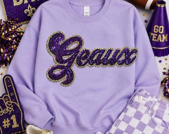 May include: A light purple crewneck sweatshirt featuring the word Geaux in purple sequins with a gold sequin border. The garment is displayed alongside a gold football helmet, a brown football, a purple megaphone, and gold glitter sneakers.