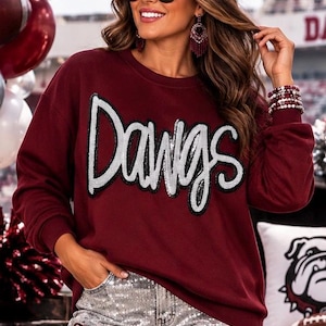 May include: A maroon long-sleeve sweatshirt featuring the word Dawgs in large, silver sequined lettering across the chest. The garment has a crew neckline and ribbed cuffs, paired with silver sequined pants that include small maroon ribbon bows.