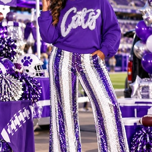 May include: A purple long-sleeve sweatshirt featuring the word Cats in silver sequined script across the chest, paired with wide-leg trousers covered in vertical stripes of purple and silver sequins and white platform block-heeled boots.