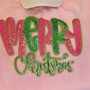 K&ouml;nnte beinhalten: Ein hellrosa Sweatshirt mit den Worten "MERRY CHRISTMAS" in Paillettenbuchstaben. Das Wort "MERRY" ist in rosa und gr&uuml;nen Pailletten, und "CHRISTMAS" ist in gr&uuml;nen Pailletten. Oben ist eine wei&szlig;e Baseballkappe zu sehen.
