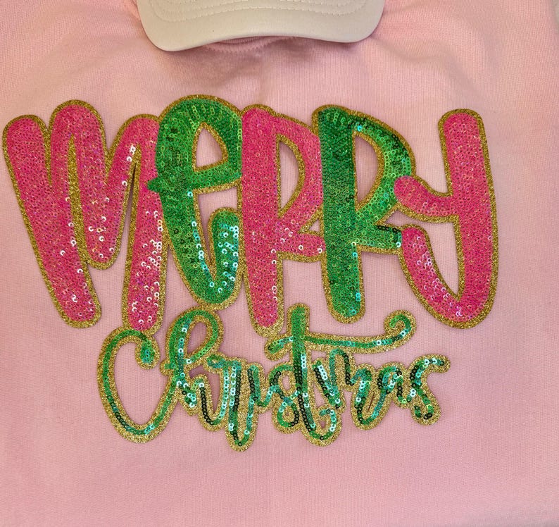 K&ouml;nnte beinhalten: Ein hellrosa Sweatshirt mit den Worten "MERRY CHRISTMAS" in Paillettenbuchstaben. Das Wort "MERRY" ist in rosa und gr&uuml;nen Pailletten, und "CHRISTMAS" ist in gr&uuml;nen Pailletten. Oben ist eine wei&szlig;e Baseballkappe zu sehen.