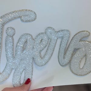 以下が含まれることがあります： 銀色のスパンコールで「Tigers」と書かれたスクリプト。この単語は筆記体で、各文字は銀色のスパンコールで縁取られています。文字は白い背景にあります。画像は装飾品です。