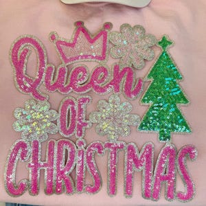 May include: A light pink sweatshirt with the words "Queen of Christmas" in pink sequined letters. The design includes a pink crown, green sequined Christmas tree, and silver sequined snowflakes. A white baseball cap is visible at the top.