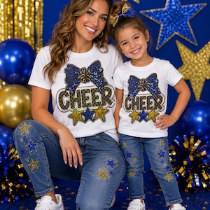 May include: Two white t-shirts featuring a blue sequined bow and the word CHEER in a checkered pattern, accented by blue and gold stars. The shirts are paired with blue denim jeans decorated with blue and gold star patches and white trainers.