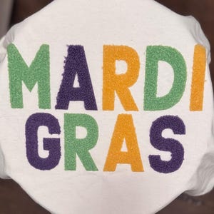 May include: White fabric featuring the words "MARDI GRAS" in large, textured letters. The letters are in green, purple, and orange, creating a vibrant design. The fabric appears to be a soft material.