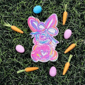 Puede incluir: Una composición con temática de Pascua sobre hierba verde. Una decoración brillante en forma de conejito rosa y azul está rodeada de zanahorias naranjas y huevos rosas y azules. El conejito tiene forma de corazón en su vientre.