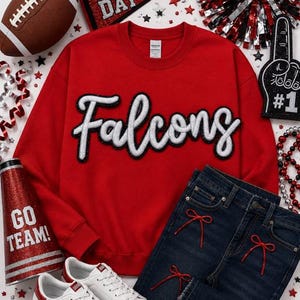 May include: A red crewneck sweatshirt featuring the word Falcons in white script with a black outline. The outfit includes dark blue denim jeans with small red ribbon bows, white sneakers with red stars, a football, a megaphone, and pom-poms.