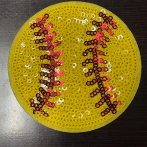 Op de afbeelding: Een ronde, gele softbal patch bedekt met pailletten. De patch heeft rode en bruine stikseldetails, die de naden van een softbal nabootsen. De pailletten geven de patch een glanzende, getextureerde uitstraling. Een leuk accessoire voor sportfans.