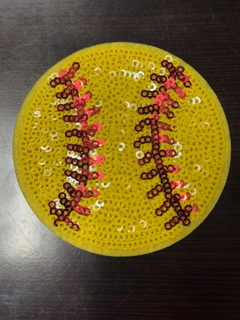 Op de afbeelding: Een ronde, gele softbal patch bedekt met pailletten. De patch heeft rode en bruine stikseldetails, die de naden van een softbal nabootsen. De pailletten geven de patch een glanzende, getextureerde uitstraling. Een leuk accessoire voor sportfans.