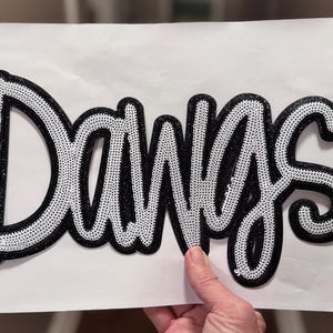 May include: A black and white sequined patch with the word "Dawgs" in a cursive font. The letters are outlined in black glitter and filled with white sequins. The patch is on a white background.