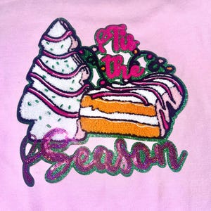 May include: Pink sweatshirt with a festive design featuring a green and white striped Christmas tree, a slice of cake with orange frosting, and the text "Tis the Season" in green and purple sequins.