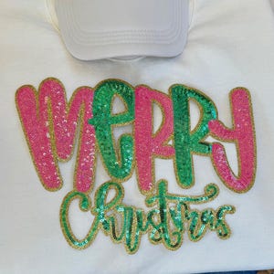 K&ouml;nnte beinhalten: Ein wei&szlig;es Sweatshirt mit dem Schriftzug "MERRY CHRISTMAS" in Paillettenbuchstaben. Das Wort "MERRY" ist in rosa und gr&uuml;nen Pailletten mit goldenem Rand gehalten. Das Wort "CHRISTMAS" ist in gr&uuml;nen Pailletten mit goldenem Rand gehalten. Eine wei&szlig;e Baseballkappe liegt obenauf.