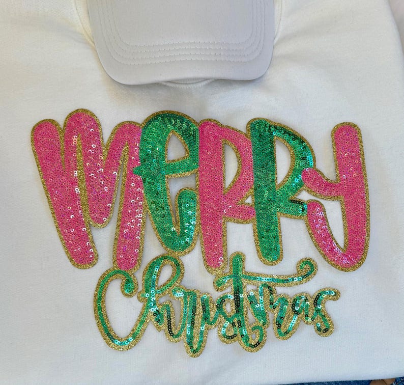 K&ouml;nnte beinhalten: Ein wei&szlig;es Sweatshirt mit dem Schriftzug "MERRY CHRISTMAS" in Paillettenbuchstaben. Das Wort "MERRY" ist in rosa und gr&uuml;nen Pailletten mit goldenem Rand gehalten. Das Wort "CHRISTMAS" ist in gr&uuml;nen Pailletten mit goldenem Rand gehalten. Eine wei&szlig;e Baseballkappe liegt obenauf.
