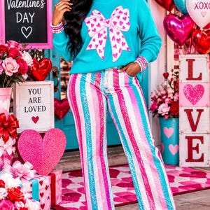 May include: A bright turquoise long-sleeve sweater featuring a large white bow graphic with pink hearts, paired with high-waisted pants covered in vertical stripes of pink, white, and turquoise sequins, finished with turquoise ankle boots.