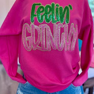 Puede incluir: Una sudadera rosa brillante con las palabras "Feelin' Grinchy" en letras de lentejuelas verdes y rosas. La sudadera de manga larga se combina con pantalones cortos de mezclilla azules. El texto es el elemento principal de la prenda.