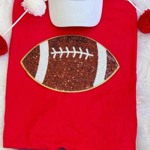 May include: A red t-shirt featuring a sequined football design with white stripes and stitching. A white baseball cap is placed above the shirt. The shirt is displayed on a white fluffy surface.