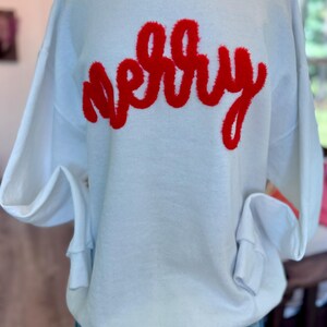 Puede incluir: Sudadera blanca con la palabra "merry" en letras rojas y esponjosas en el pecho. La parte superior de manga larga está hecha de un material suave y es adecuada para ropa informal. Las mangas tienen puños en las muñecas.