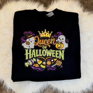 May include: Black sweatshirt with a colorful sequined design. The design features the text "Queen of Halloween" with a gold crown, ghosts, and Halloween candy. The sweatshirt is on a white fluffy surface.