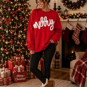 May include: A bright red long-sleeve sweatshirt featuring the word merry in white fuzzy script lettering across the chest, paired with solid black leggings and tan suede ankle boots with a white faux fur lining at the top edge.