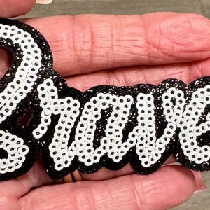 May include: A decorative patch featuring the word Braves in white sequins outlined with a black glitter border. The script lettering is connected and has a raised, dimensional appearance. The design is presented against a dark, sparkling background.