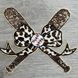 Puede incluir: Un bate de béisbol de lentejuelas marrón y dorado y un lazo de estampado de leopardo con una pelota de béisbol blanca en el centro. El lazo está atado alrededor de los bates.