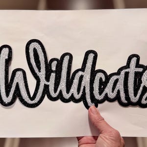 May include: A black and white Wildcats iron-on patch. The word "Wildcats" is written in a cursive font with a black glitter outline and white dotted fill. The patch is held by a hand against a white background.