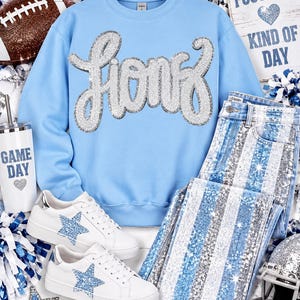 Puede incluir: Una sudadera azul claro de cuello redondo con la palabra Lions en lentejuelas plateadas, combinada con zapatillas blancas con estrellas de purpurina azul y vaqueros a rayas azules y plateadas. Incluye un vaso blanco con la etiqueta Game Day.