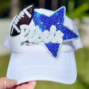 Puede incluir: Gorra de camionero blanca con un balón de fútbol y una estrella de lentejuelas azules. La palabra "Rebels" está escrita con lentejuelas blancas. La gorra tiene una parte trasera de malla y una visera curva.