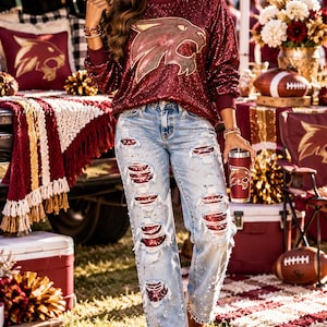 May include: A long-sleeve maroon sequin sweatshirt featuring a gold and maroon wildcat mascot graphic, paired with light-wash distressed denim jeans that have maroon sequin patches visible through the rips and a maroon tumbler with a wildcat logo.