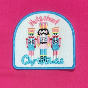 Puede incluir: Un parche bordado blanco y azul con tres figuras de cascanueces y el texto "Nuts about Christmas".