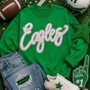 May include: A bright green crewneck sweatshirt featuring the word Eagles in large white sequined script lettering across the chest. The garment is paired with light blue distressed denim jeans with green sequin patches and white sneakers.