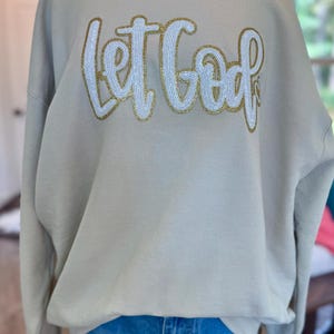 May include: A beige sweatshirt with the phrase "Let God" in white sequined letters outlined in gold. The sweatshirt is displayed on a mannequin, paired with blue denim shorts.
