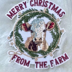 May include: Embroidered patch with a cow's face inside a green wreath, with the text "MERRY CHRISTMAS FROM THE FARM" in burgundy. The cow has a light blue and brown coat, with red accents on the ears. The patch is on a white background.