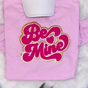 Op de afbeelding: Een lichtroze t-shirt met de woorden "Be Mine" in een retro lettertype, omlijnd met gouden glitter. Een witte baseballpet ligt bovenop het shirt. Het shirt heeft korte mouwen met een ruchedetail.