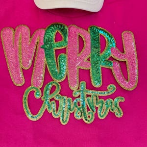 K&ouml;nnte beinhalten: Ein pinkfarbenes T-Shirt mit dem Schriftzug "MERRY CHRISTMAS" in Paillettenbuchstaben. Das Wort "MERRY" ist in pinkfarbenen Pailletten mit goldenem Rand, die Buchstaben "M" und "R" sind gr&uuml;n. Das Wort "CHRISTMAS" ist in gr&uuml;nen Pailletten mit goldenem Rand.