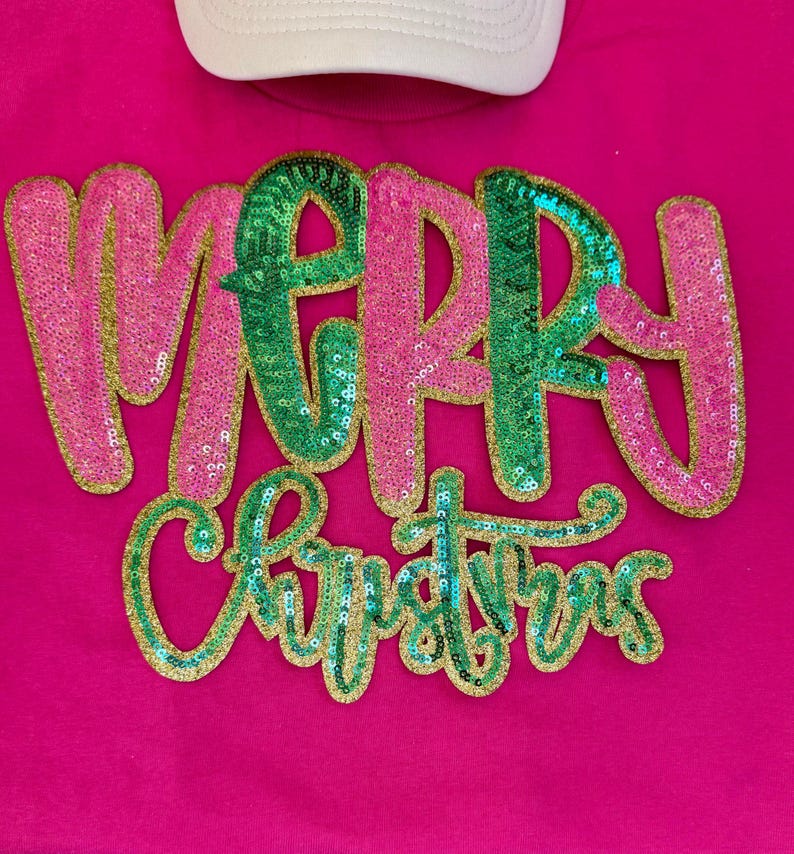 K&ouml;nnte beinhalten: Ein pinkfarbenes T-Shirt mit dem Schriftzug "MERRY CHRISTMAS" in Paillettenbuchstaben. Das Wort "MERRY" ist in pinkfarbenen Pailletten mit goldenem Rand, die Buchstaben "M" und "R" sind gr&uuml;n. Das Wort "CHRISTMAS" ist in gr&uuml;nen Pailletten mit goldenem Rand.