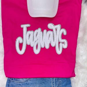 May include: A hot pink t-shirt with the word "Jaguars" in white sequins outlined in silver. A white baseball cap is positioned above the shirt. The bottom of the shirt is paired with blue denim jeans.