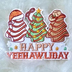 May include: Embroidered patch with a red and white Santa, a green Christmas tree with gold stars, and a cowboy with a brown hat, all wrapped in red and white ribbon. The text "HAPPY YEEHAWLDIDAY" is below the figures.