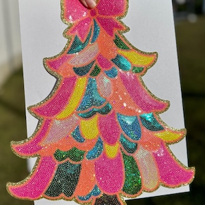 May include: A vibrant, sequined Christmas tree decoration. The tree has a pink bow at the top and is decorated with sequins in pink, orange, yellow, teal, and green. The edges are outlined with gold glitter.