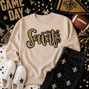 Peut inclure: Un sweat-shirt à col rond beige avec le mot Saints écrit en lettres cursives à paillettes dorées et contour noir. Le vêtement est associé à un jean noir à motif léopard discret et détails fleur de lys dorés, ainsi qu'à des baskets blanches.