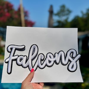 Könnte beinhalten: Ein weißer Schriftzug-Ausschnitt mit dem Wort Falcons, bestehend aus dicken, erhabenen weißen Buchstaben mit einer schwarzen Glitzerumrandung, präsentiert auf einem schlichten weißen rechteckigen Hintergrund im Freien.