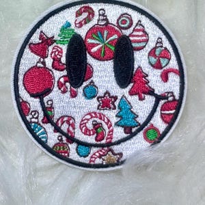 May include: White embroidered iron-on patch with a smiling face surrounded by Christmas-themed designs, including candy canes, ornaments, and trees.