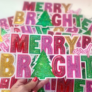 May include: A close-up of a stack of colorful, sparkly iron-on patches that say "Merry and Bright" with a green sequined Christmas tree in the center.