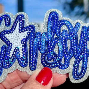 May include: A sequined patch in the shape of the word "Cowboys" in blue script with a white star. The patch has a silver glitter border and is held by a hand with red nail polish.