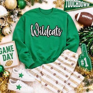 Puede incluir: Una sudadera verde de cuello redondo con la palabra Wildcats en letras cursivas negras y blancas, combinada con pantalones blancos con rayas de purpurina dorada e iconos de balones de fútbol, y zapatillas blancas con estrellas verdes.