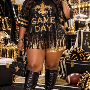 May include: A black sequined t-shirt with gold fringe, a gold fleur-de-lis emblem, and the text "GAME DAY" in gold lettering. The shirt is paired with gold sequined shorts, black knee-high boots, and a black quilted handbag with a gold chain.
