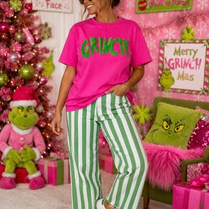 Op de afbeelding: Een felroze T-shirt met korte mouwen en het woord GRINCH in pluizige groene letters op de borst, gecombineerd met een wit-groene verticaal gestreepte spijkerbroek met een rafelige zoom aan de onderkant van de broekspijpen.